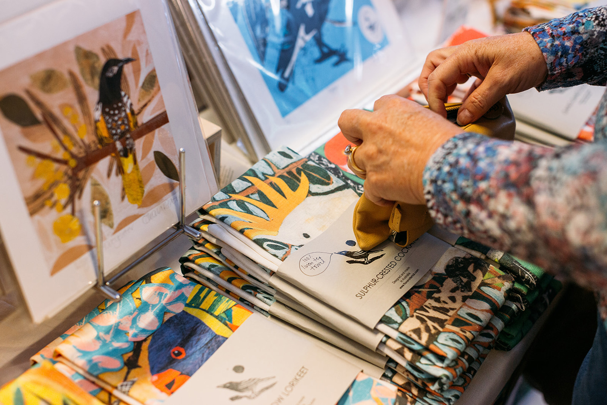 Two female hands looking through a selection of printed artworks and textiles at a market stall.