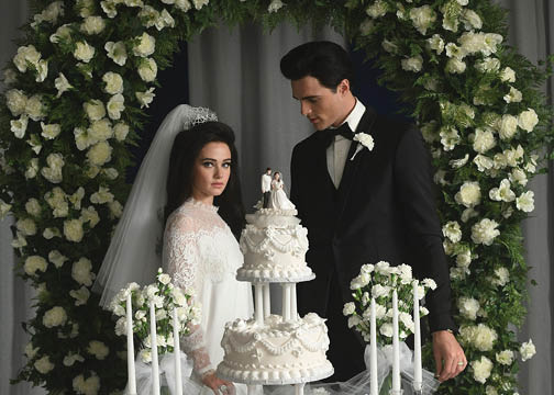 Still from film Priscilla. Characters of Elvis and Priscilla standing behind their wedding cake under an arch of white flowers.