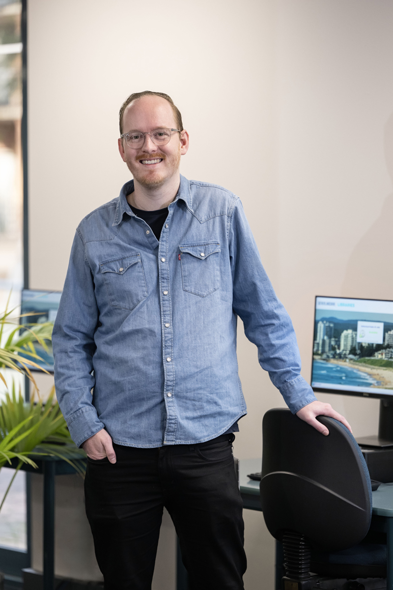 Man with glasses standing beside a desk with a computer on it.