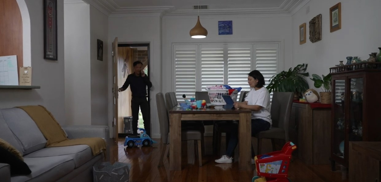 A woman sitting at a dining room table on a laptop while her husband enters the room