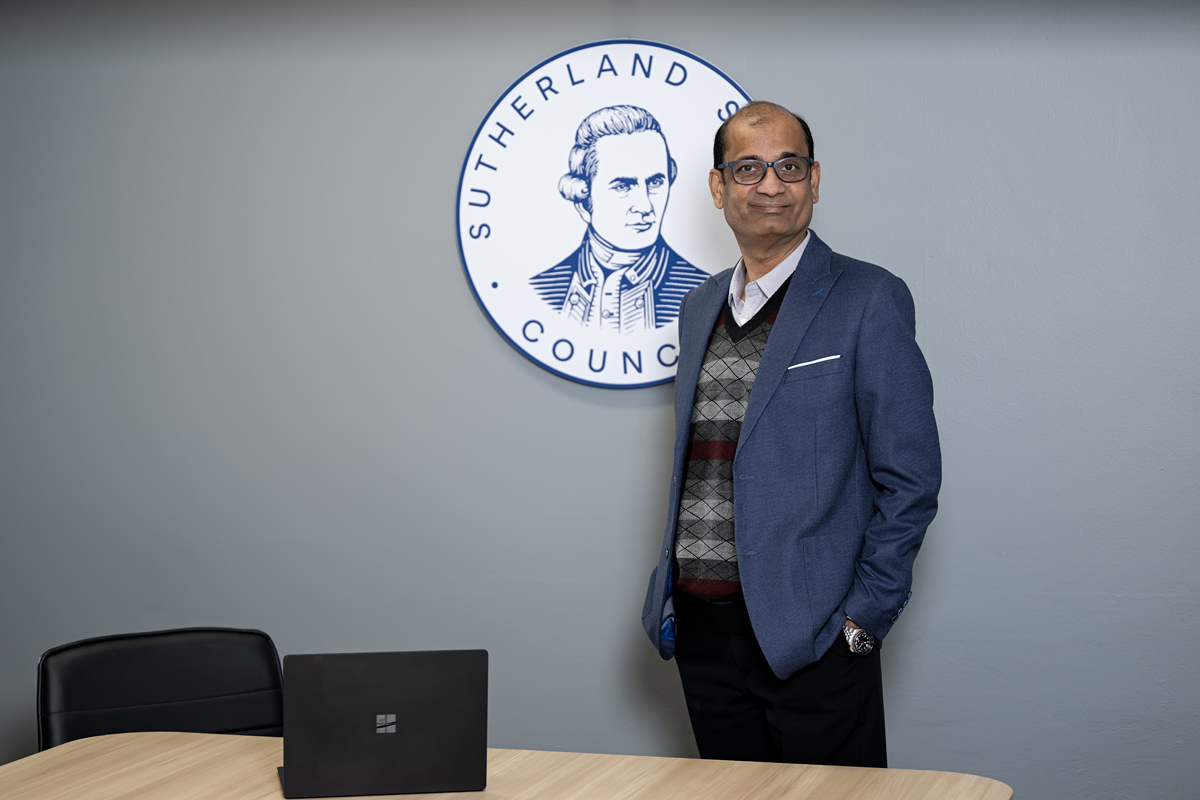 man in business attire standing in an office with a computer on the desk in front of him. 