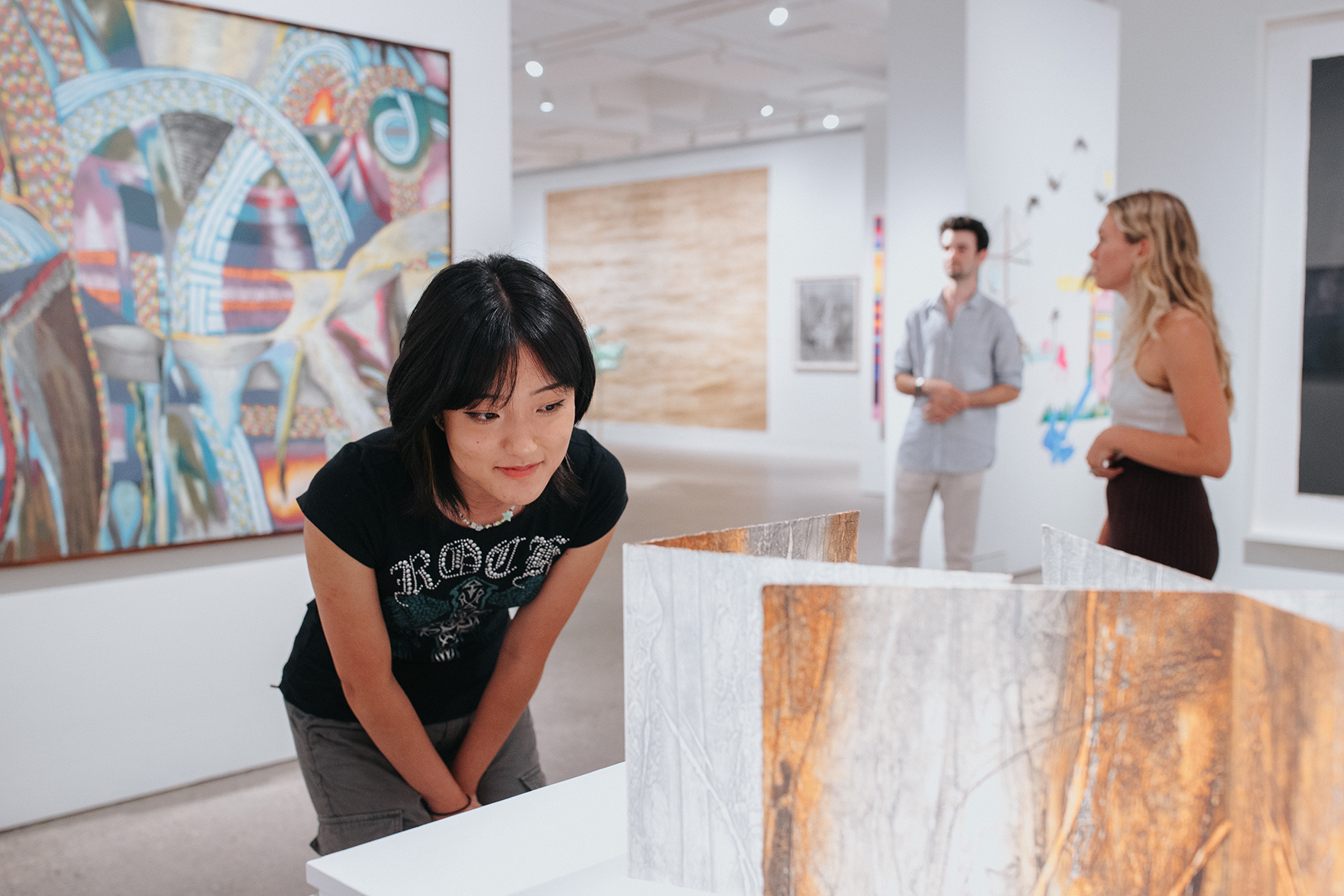 Young woman in gallery space bending to look at a sculptural work on a plinth.