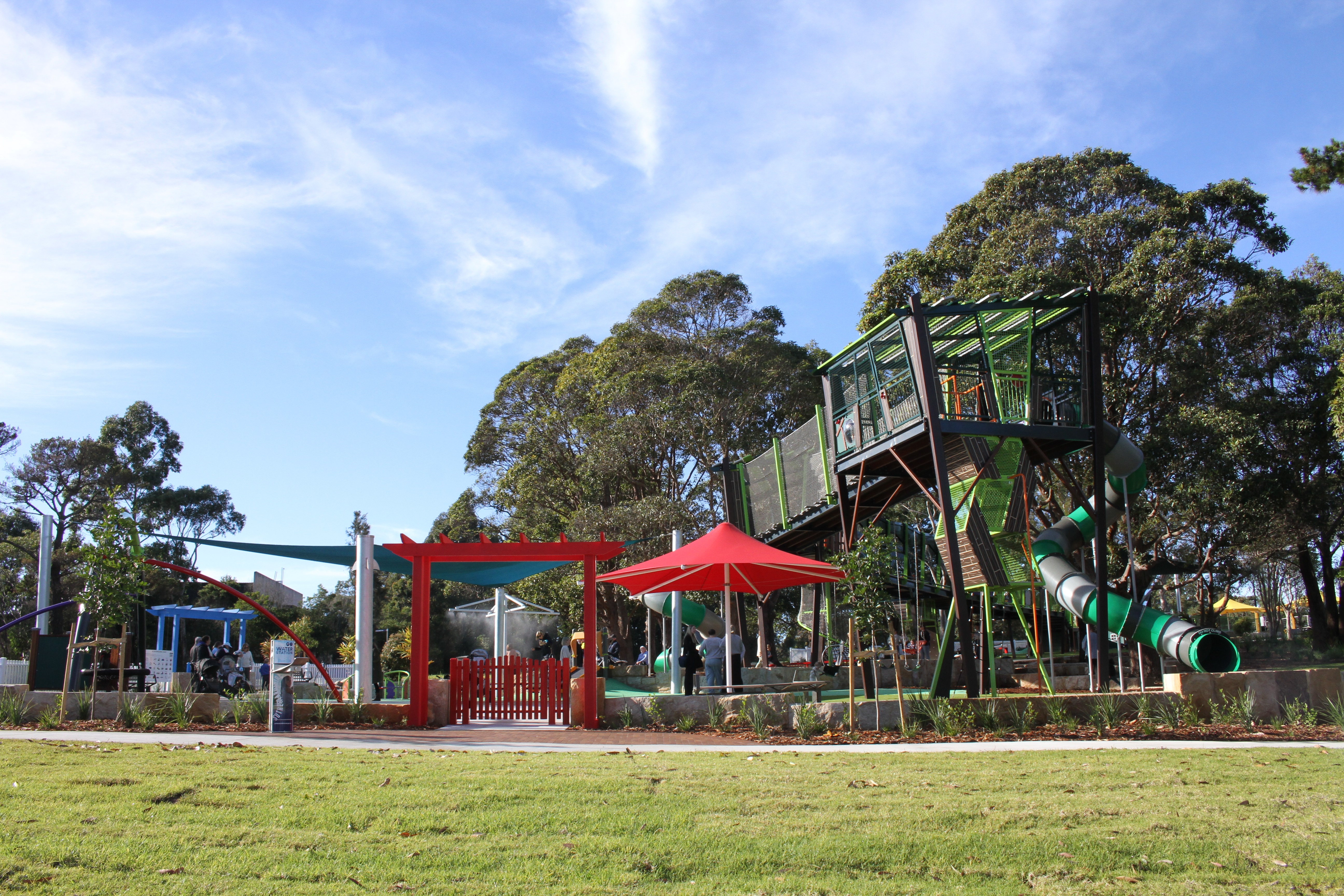 A playground with slide, shading and swings. Trees and blue sky in the background, green grass and footpath in the foreground