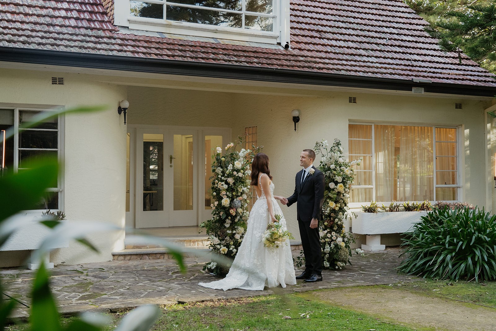 A man and woman in bridal attire posing in front of the Hazelhurst cottage, an art deco cottage painted pale yellow with white trim.