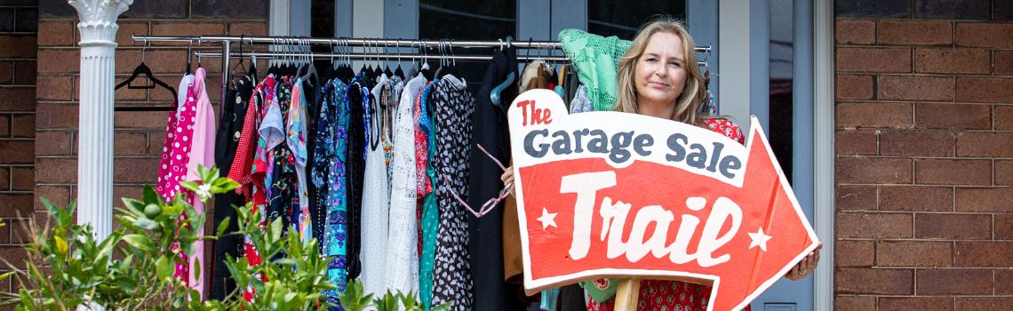 A woman standing over a clothing rack smiling, speech bubble that says Buy, Sell, Reuse. Garage Sale Trail logo