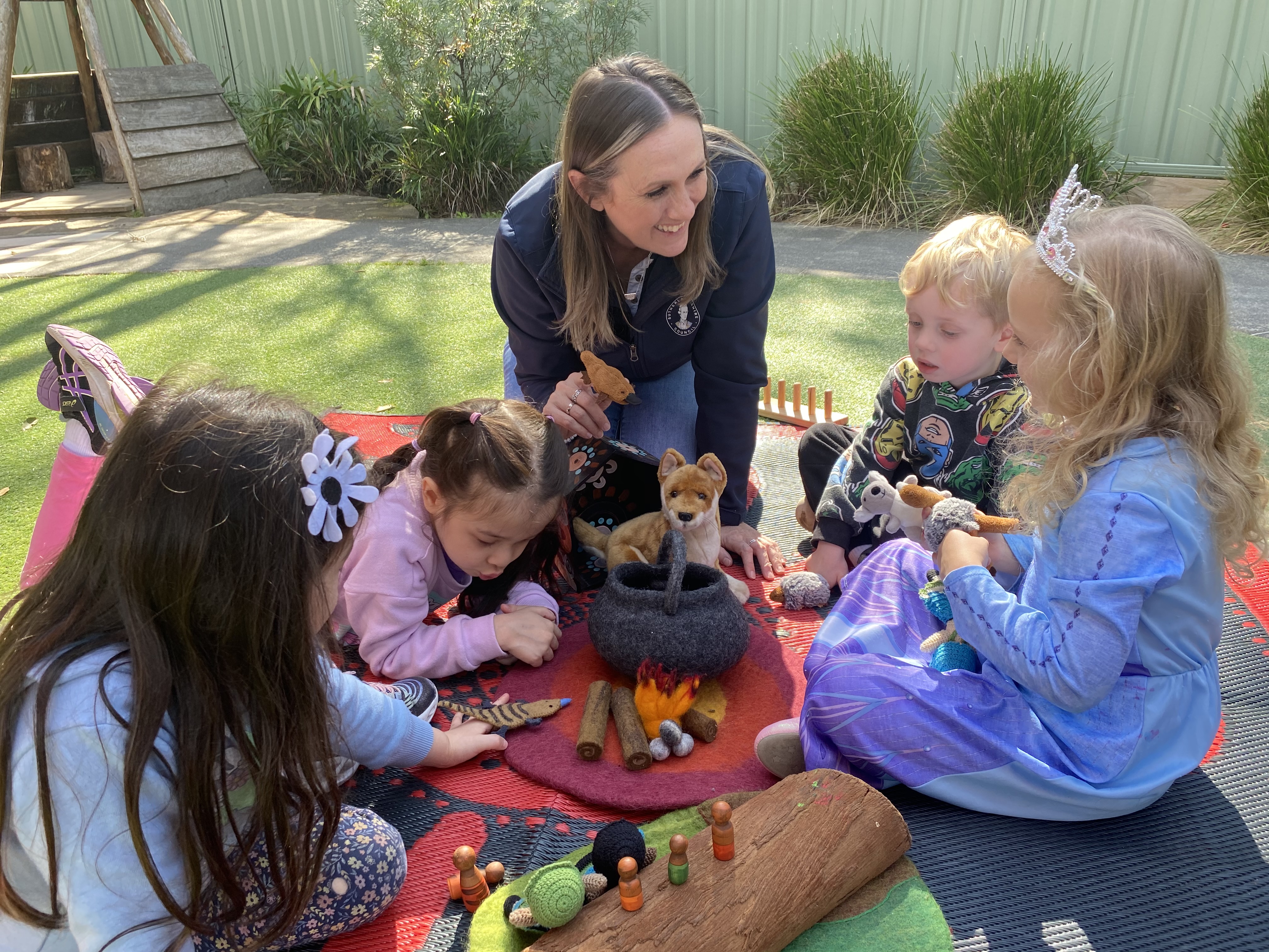 An Early Education Centre Educator sitting and playing with a group of children on a colourful rug