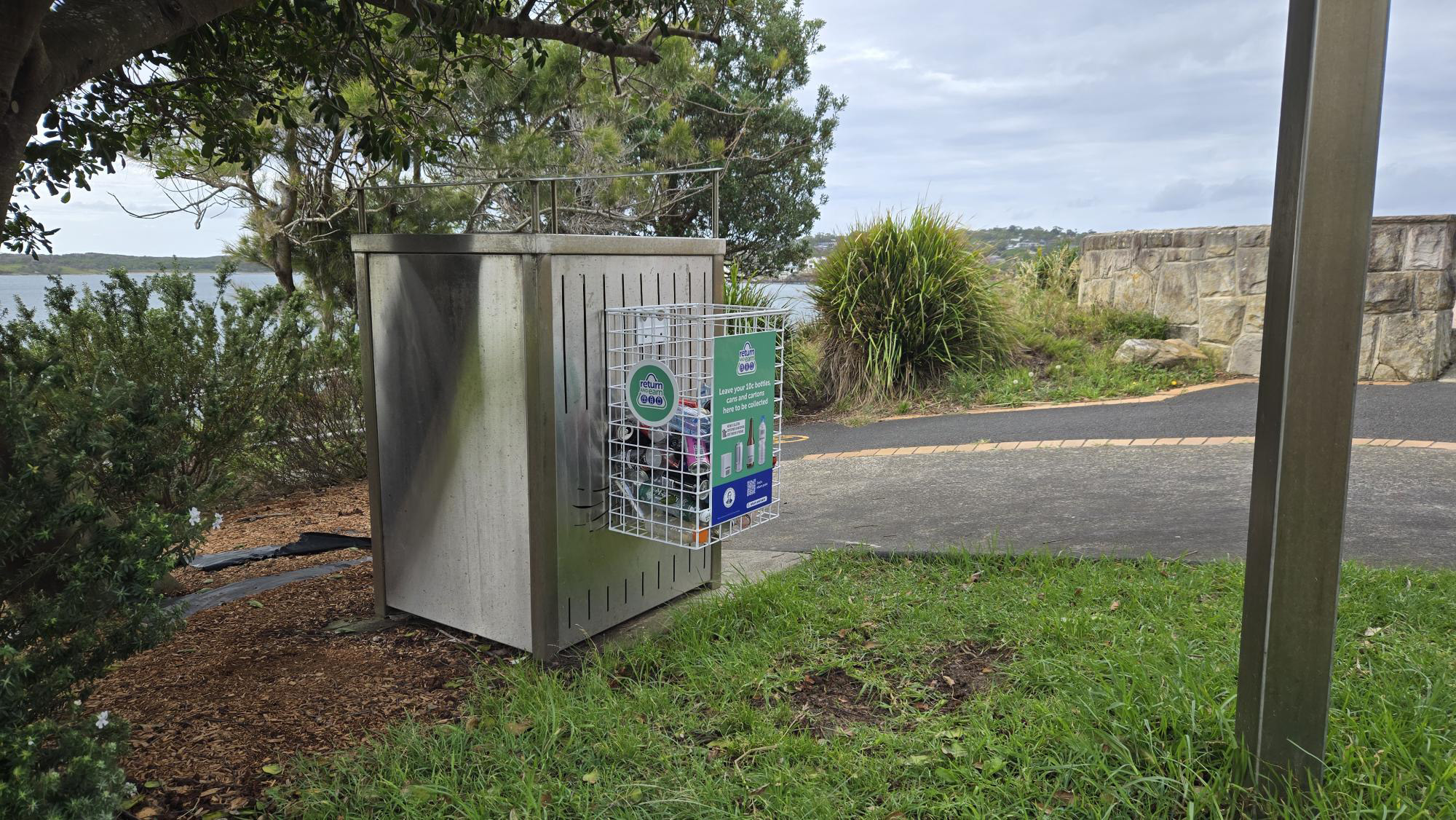 A rubbish bin with a return and earn basket attached full of cans and bottles. Pathway, gardens and water in background