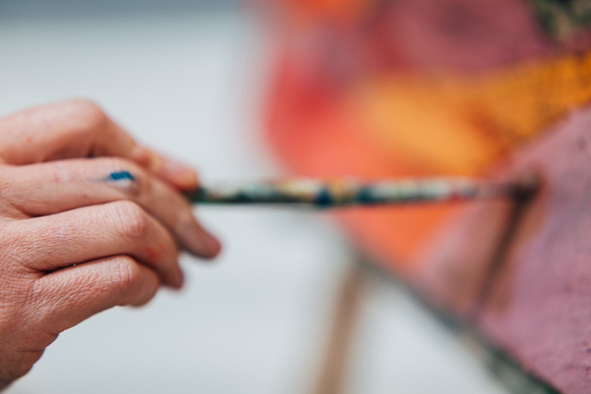 Close up photograph of a hand holding a paintbrush in front of a brightly coloured canvas.