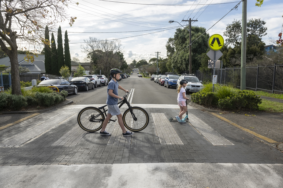 Boy walking bike and Girl walking scooter across a pedestrian crossing.